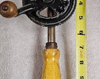 Puede incluir: Taladro manual vintage con mango negro y amarillo, engranaje negro y mango de madera. El taladro mide aproximadamente 30 cm de largo. Una cinta métrica amarilla se coloca junto al taladro.