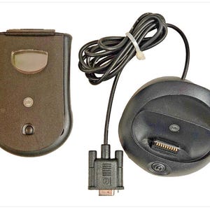 May include: A black electronic device with a small screen and a round button, next to a charging dock and a cable with a connector. The charging dock has a slot for the device and a round button. The items are on a white background.