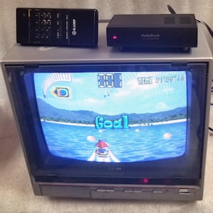 May include: A silver CRT television displaying a jet ski video game with the text Goal on the screen. A black Kamp remote control and a black RadioShack RF modulator with a red light are placed on top of the television set, which has front buttons.