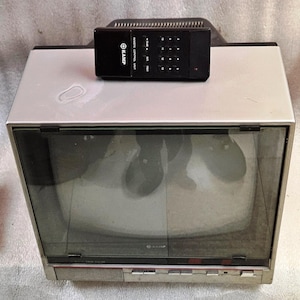 May include: A silver and black vintage television set with a glass screen and a matching black remote control unit labelled KAMP resting on top. The television features a front-facing speaker grille, several control buttons, and a dark screen.