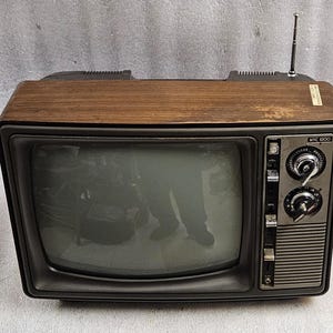 May include: A vintage NTC 1900 television set featuring a wood-grain top cabinet, a black plastic front frame, and a silver control panel with two large circular knobs. A thin metal aerial extends upwards from the top right corner of the unit.