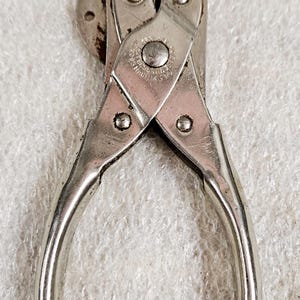 May include: A pair of silver-toned metal fencing pliers with a pointed jaw, a side-mounted wire cutter, and a central pivot point. The tool features visible rivets and stamped text on the metal surface that reads 'MADE IN JAPAN'.