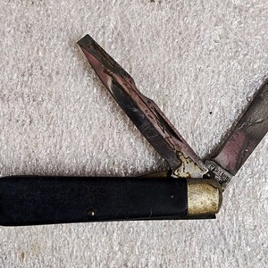 May include: A vintage pocket knife with two blades open. The blades show signs of wear and discoloration. The handle is black with a metal end and a small loop for attaching to a keychain.
