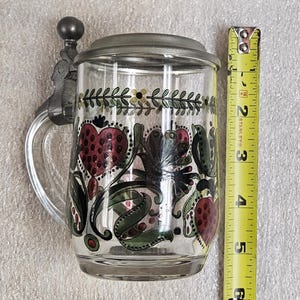 May include: Clear glass beer stein with a hinged metal lid and handle. The stein is decorated with a floral design featuring red hearts and green leaves. The stein measures approximately 5 inches tall.