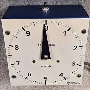 Może przedstawiać: Zegarek stopera w stylu vintage z białą tarczą i niebieskim wierzchem. Tarcza zegara wyświetla liczby od 1 do 10, ze słowem "seconds" pod centralną wskazówką. Marka "DAMON" jest wydrukowana na dole.