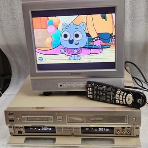 May include: A silver Sharp TV displaying a cartoon with a blue character. Below the TV is a JVC VCR with a remote. The VCR has a Super VHS ET logo and a digital display.