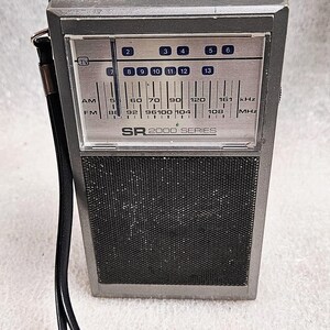 May include: A vintage silver and black SR 2000 SERIES radio with a telescopic aerial. The radio features a black speaker grille and a tuning dial with AM/FM frequencies. A black strap is attached to the side.