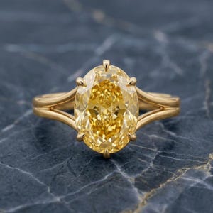 Könnte beinhalten: Ein goldener Ring mit einem großen, oval geschliffenen gelben Diamanten. Der Ring hat ein geteiltes Schaftdesign und vier Krappen, die den Edelstein halten. Die Facetten des Diamanten reflektieren das Licht und erzeugen ein brillantes Funkeln. Der Ring ist vor einem dunklen, strukturierten Hintergrund platziert.