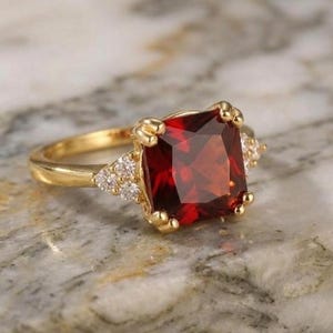 May include: A gold ring with a large, square-cut, deep red gemstone. The ring has a classic design with small, clear accent stones on either side of the main stone. The band is a polished gold colour, and the ring is set against a marble surface.