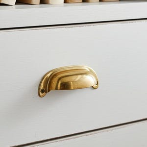 May include: A white painted wooden cabinet with a brass drawer pull. The top shelf displays a collection of vintage books with various spines and titles. A small white jar is also visible on the shelf.