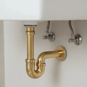 Op de afbeelding: Close-up van sanitair onder een witte wastafel. Zichtbaar zijn een goudkleurige P-val, verticale buizen en zilveren afsluiters tegen een witte muur. De buizen hebben een glanzende, metalen afwerking.