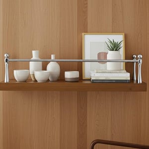 Puede incluir: Una estantería de madera con una barra cromada sostiene jarrones, cuencos y una pequeña planta en maceta de cerámica blanca. Una impresión artística enmarcada y libros también están en la estantería. La estantería está montada en una pared de madera.