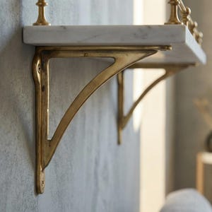 Puede incluir: Una estantería de mármol con soportes de latón. La estantería es blanca con vetas grises. Los soportes son de color dorado cálido y están montados en una pared gris. El diseño es elegante y adecuado para la decoración del hogar.