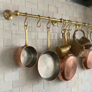 Op de afbeelding: Een messing keukenrek met hangende koperen potten en pannen. Het rek is bevestigd aan een witte betegelde muur. Diverse keukengerei en een kleine geweven mand hangen ook.