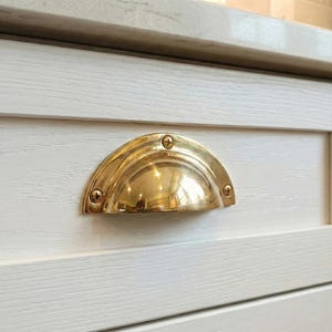 May include: Close-up of a brass-colored, half-moon drawer pull mounted on a white wooden cabinet. The pull is secured with four small screws and reflects light. The cabinet has a light-colored countertop.