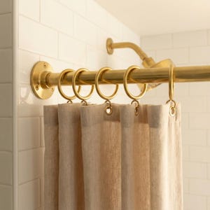 May include: A gold-colored shower curtain rod with matching rings and a beige linen shower curtain. The rod is mounted on a white tiled wall, creating a clean and elegant bathroom aesthetic.