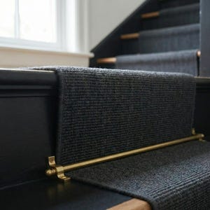 May include: Close-up of a dark gray stair runner with a brass rod. The runner is installed on a black painted staircase. The brass rod is mounted on the side of the stairs.