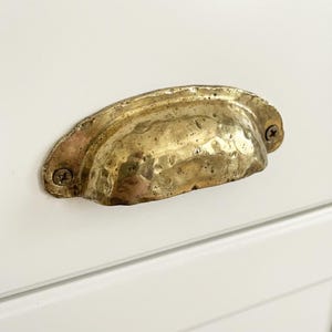 Puede incluir: Un tirador de cajón de latón martillado, con forma de copa, montado sobre una superficie blanca con dos tornillos. El latón tiene un aspecto cálido y envejecido. Para muebles.