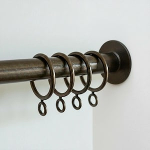 May include: Bronze-colored curtain rod with matching rings and end cap. The rod is mounted on a white wall. The rings are designed to hold curtains and slide along the rod. The end cap is circular and provides a finished look.