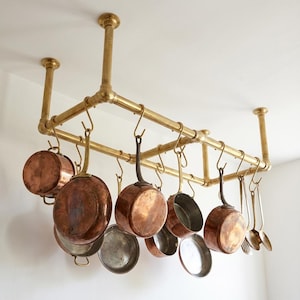 May include: A brass-colored pot rack suspended from the ceiling, holding a collection of copper pots and pans. The rack is made of brass pipes and features multiple hooks for hanging cookware. The pots and pans vary in size and shape, with copper exteriors.