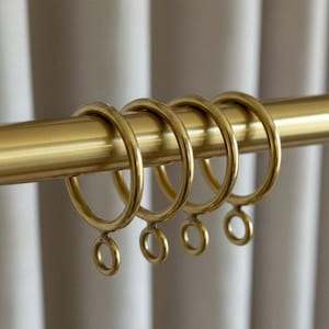 May include: Close-up of a gold-colored curtain rod with matching rings. The rings are circular with small loops at the bottom for attaching curtains. The rod and rings have a shiny, metallic finish. The background is a soft, neutral color.