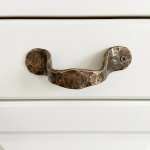 Op de afbeelding: Een bronzen, gehamerde metalen trekker met een gebogen handvat. De trekker is met twee schroeven aan een witte lade voorkant bevestigd. Het metaal heeft een getextureerd oppervlak en een rustieke uitstraling.