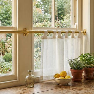 May include: A kitchen window featuring a brass curtain rail and cream linen curtains. A clear glass bottle, a bowl of lemons, and potted herbs are on the wooden windowsill. The window offers a view of a green garden.