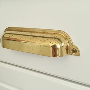 Könnte beinhalten: Nahaufnahme eines Messing-Schubladengriffs mit geschwungenem Design. Der Griff ist auf einer weißen Oberfläche montiert und mit zwei Schrauben befestigt. Das Messing hat ein leicht gealtertes Aussehen.