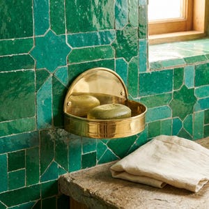May include: A brass soap dish fixed to a wall of green tiles. The dish has a semi-circular back and holds two green soap bars. A folded white towel sits on a stone surface below. The tiles are a geometric pattern.