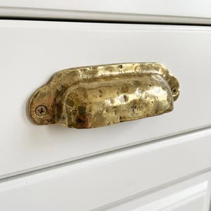 May include: A hammered brass drawer pull with a cup-shaped design. The hardware is mounted on a white cabinet door. The brass has a textured surface and is secured with two screws, showcasing a vintage aesthetic.