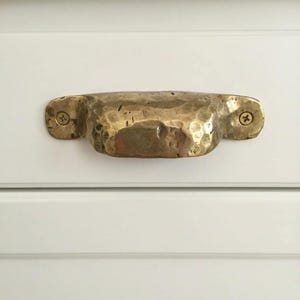 May include: A hammered brass drawer pull with a textured surface, mounted on a white cabinet door. The brass handle is secured with two screws, showcasing a rustic, handcrafted aesthetic. This hardware adds a touch of vintage charm.
