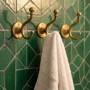 May include: Three brass-colored wall hooks with a curved design are mounted on a green geometric tile wall. A white towel hangs from one of the hooks. The hooks have a round base and a decorative ball at the top.