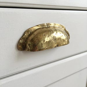 Puede incluir: Un tirador de cajón de latón martillado en forma de media luna está montado en un mueble pintado de blanco. El herraje tiene una superficie texturizada y un tono dorado cálido, que añade un toque de encanto vintage al mueble.