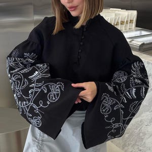 May include: A black blouse with white floral embroidery on the sleeves and a light blue skirt. The blouse has a high neckline and button details. The skirt has drawstring details. The person is wearing sunglasses and yellow heels.