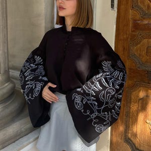 May include: A black blouse with wide sleeves featuring white floral embroidery. The blouse is paired with a light gray skirt. The image is taken outdoors in front of a wooden door.
