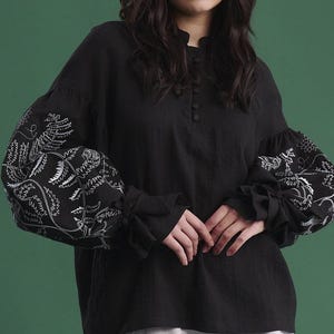 May include: Black long-sleeve blouse with white floral embroidery on the sleeves. The blouse has a high neckline and button details. Paired with a light gray mini skirt. The background is a solid green.