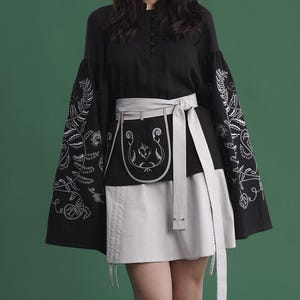 May include: Black top with embroidered sleeves and a matching skirt. The sleeves feature white floral embroidery. A gray apron-style belt with a decorative design and a matching tie cinches the waist. The outfit is set against a green backdrop.