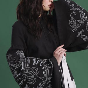 May include: Black long-sleeved top with white floral embroidery on the sleeves. The top has a high collar and button details. A white sash is tied around the waist. The background is a solid green.