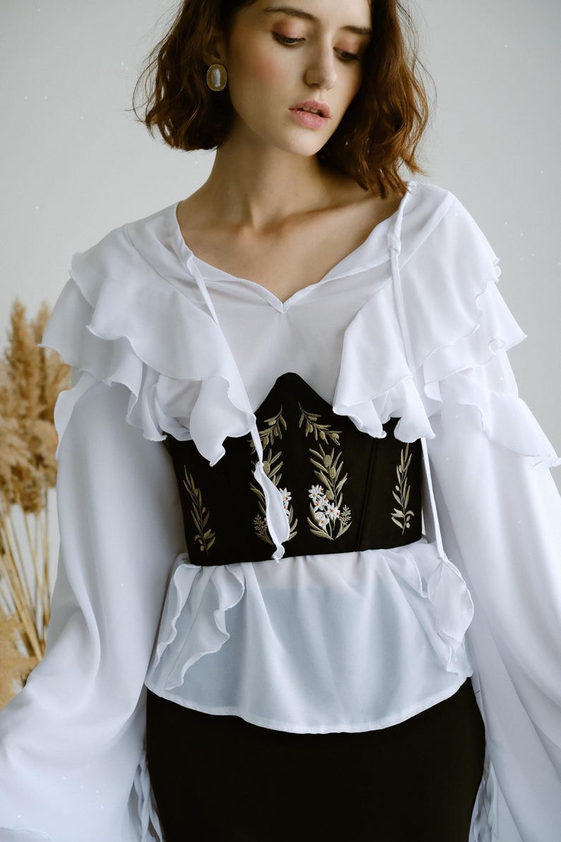 May include: A white long-sleeved blouse with layered ruffles on the shoulders and chest, paired with a black corset-style waist belt featuring embroidered green leaf patterns and small white floral details on the front center panel.