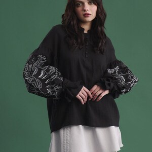 May include: Black long-sleeved blouse with white floral embroidery on the sleeves. The blouse has a high neckline and button details. Paired with a light gray skirt. The background is a solid green.