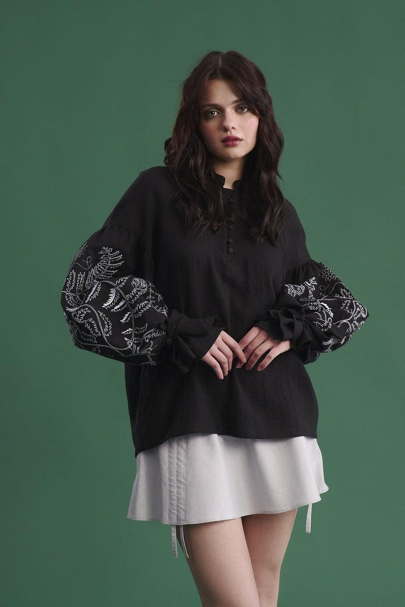 May include: Black long-sleeved blouse with white floral embroidery on the sleeves. The blouse has a high neckline and button details. Paired with a light gray skirt. The background is a solid green.
