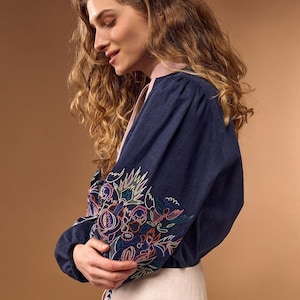 May include: A navy blue long-sleeved blouse featuring a light pink collar and cuffs, adorned with intricate multi-colored floral embroidery on the sleeves. The blouse is paired with a light beige skirt that has a vertical dark blue stitched detail.