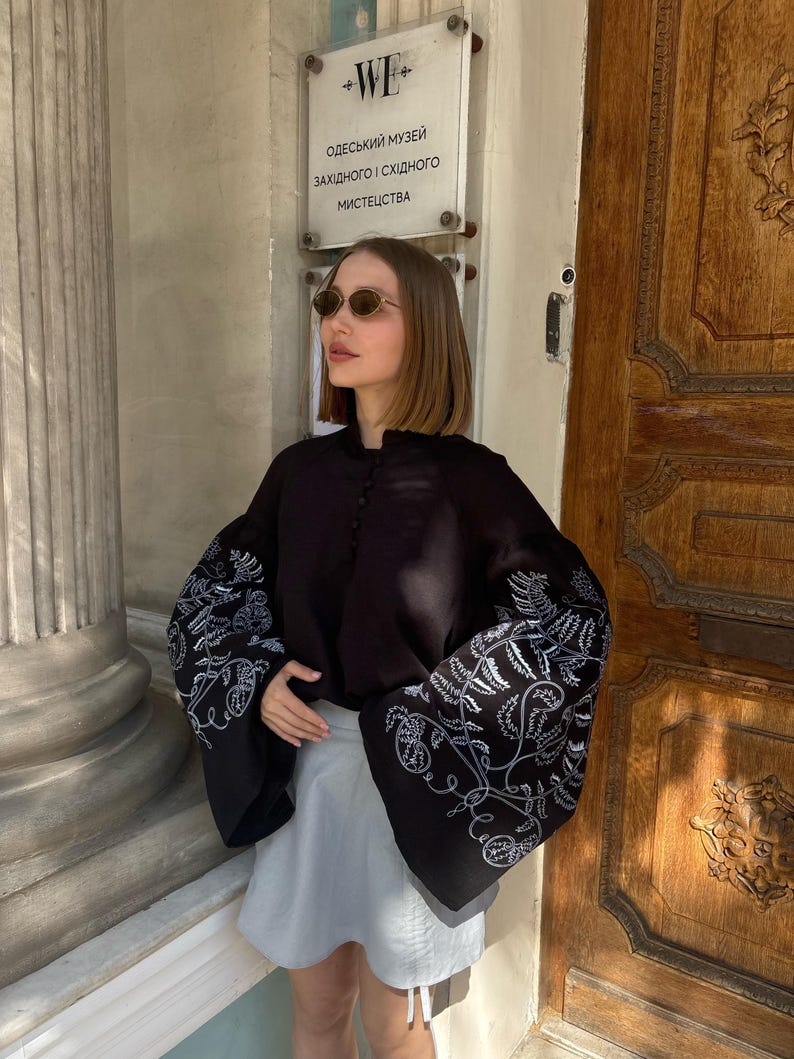 May include: A black blouse with wide sleeves featuring white floral embroidery. The blouse is paired with a light gray skirt. The image is taken outdoors in front of a wooden door.