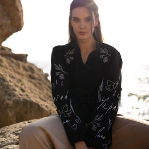 May include: A black long-sleeved shirt featuring white line-art embroidery of floral sprigs, doves, and Cyrillic text. The shirt has a V-neckline and gathered cuffs, paired with loose-fitting tan trousers, creating a contrast between the dark top and light pants.