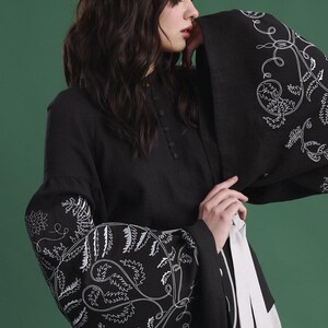 May include: Black long-sleeved tunic with white floral embroidery on the sleeves. The tunic has a high collar and button details. A white sash is tied around the waist. The background is a solid green.