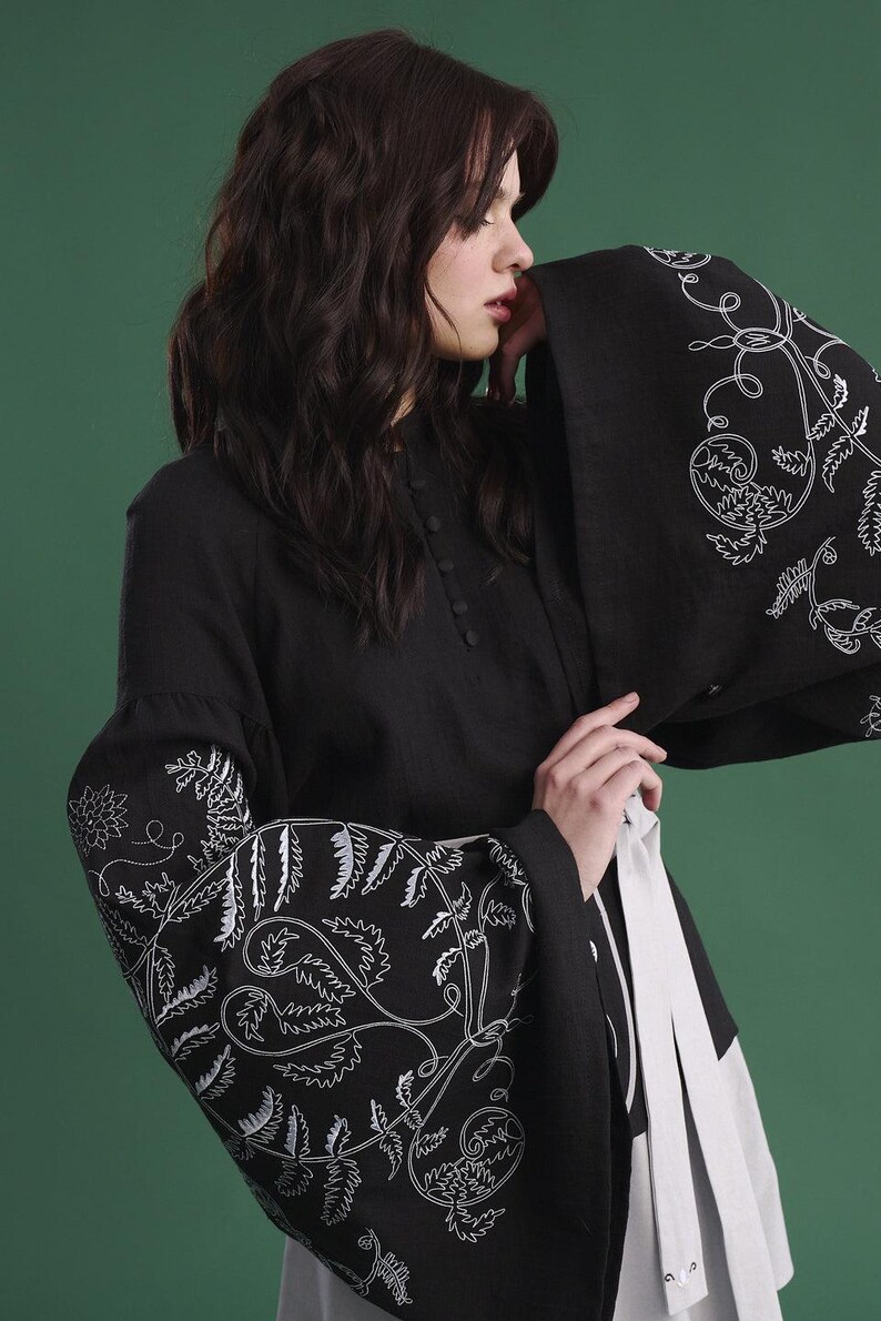 May include: Black long-sleeved tunic with white floral embroidery on the sleeves. The tunic has a high collar and button details. A white sash is tied around the waist. The background is a solid green.