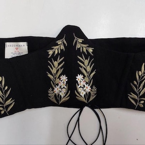 May include: A black corset top featuring embroidered olive branches with green leaves and small white flowers. The garment has a pointed center front, a white interior label with the text KROSHENKA, and thin black laces hanging from the bottom edge.