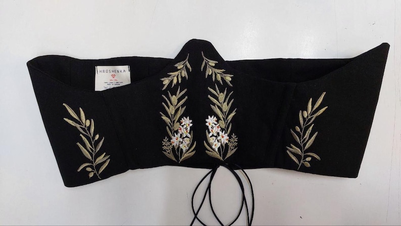 May include: A black corset top featuring embroidered olive branches with green leaves and small white flowers. The garment has a pointed center front, a white interior label with the text KROSHENKA, and thin black laces hanging from the bottom edge.
