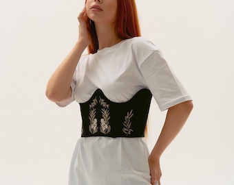 May include: A black corset-style waist belt featuring intricate light-colored floral and leaf embroidery, worn over a plain white short-sleeved t-shirt and light blue denim jeans. The corset has a structured, curved hemline and a fitted silhouette.