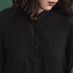 May include: Close-up of a black blouse with a mandarin collar and a series of four buttons down the front. The sleeves are long and slightly flared. The fabric has a subtle texture. The blouse is a solid black color.
